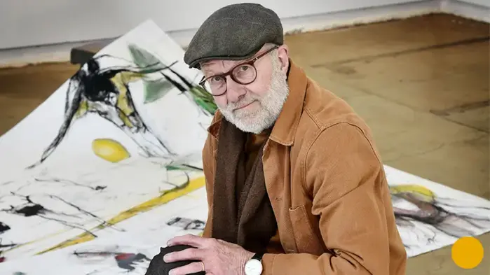Ett fotografi på Bill Olsson, han har grått skägg, glasögon och keps. I bakgrunden syns målningar.