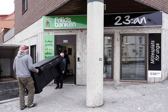 Två personer bär in en svart soffa via entrén till Fritidsbanken och 23:ans mötesplats.