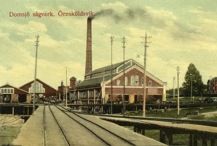 Bilden visar en röd industribyggnad med en rökande skorsten. Framför byggnaden går det tågrälser, foto 